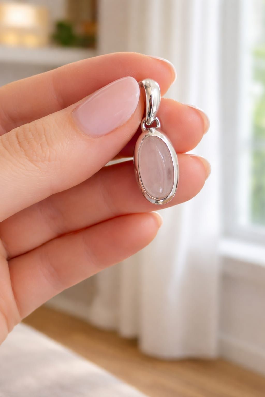 Una mano sostiene un pequeño colgante de plata con una piedra ovalada de color rosa pálido cerca de una ventana luminosa.
