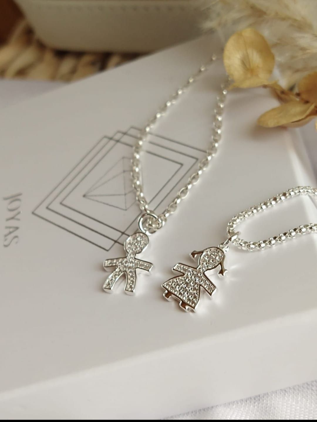 Dos collares con cruz de plata en una caja blanca con logotipo geométrico.