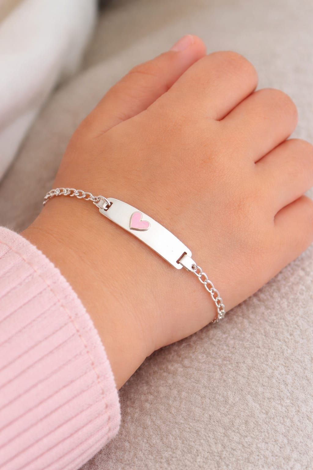 Mano que lleva una pulsera plateada con un dije de corazón rosa sobre una manga rosa suave.