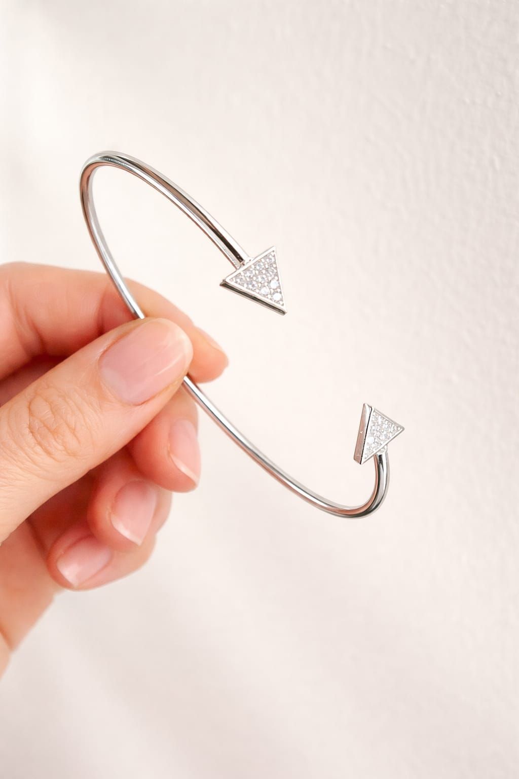 Mano sosteniendo una pulsera plateada ajustable de puño abierto con dos extremos en forma de flecha sobre un fondo blanco.