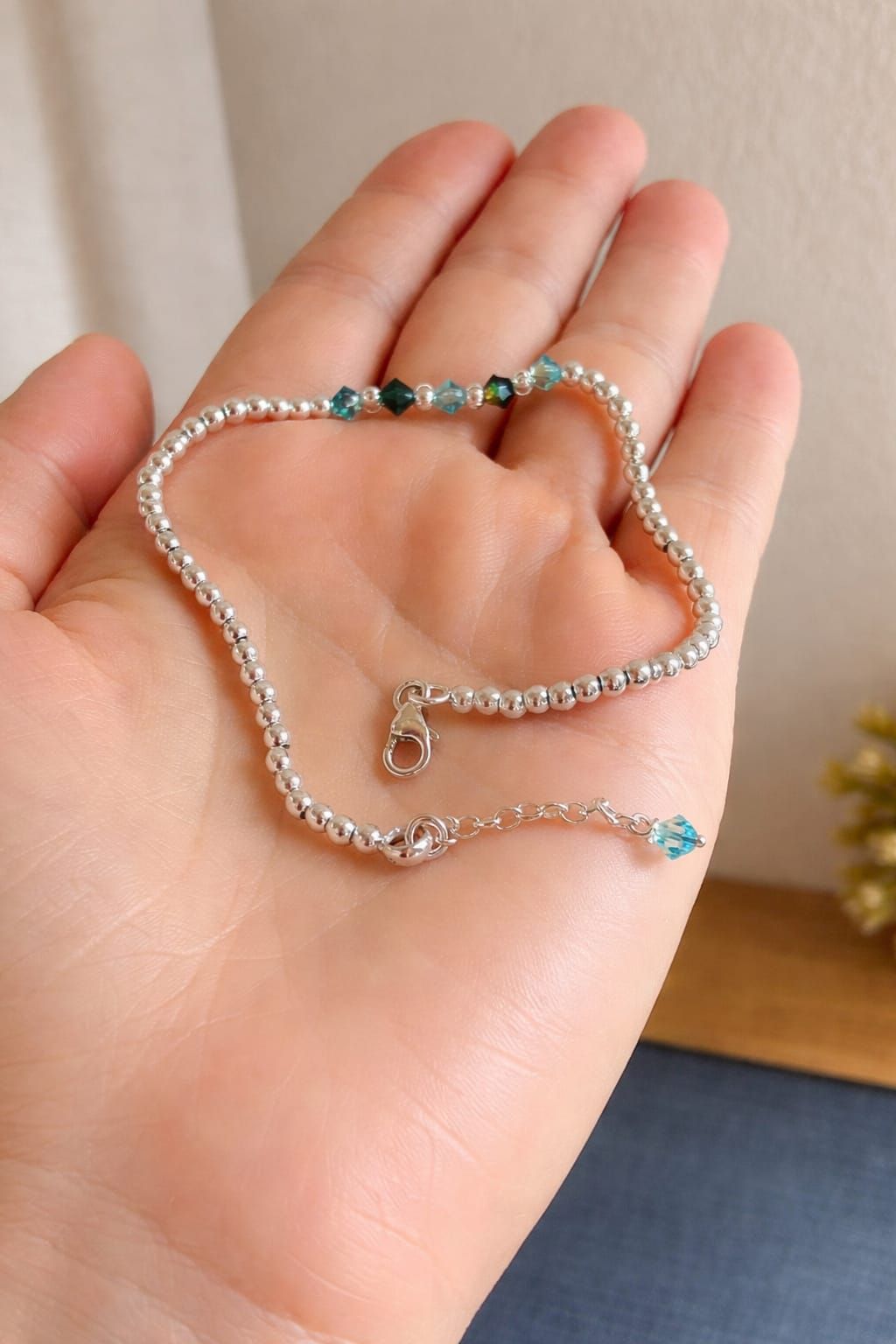 Mano que sostiene una delicada pulsera de cuentas de plata con detalles de turquesa y un pequeño dije.