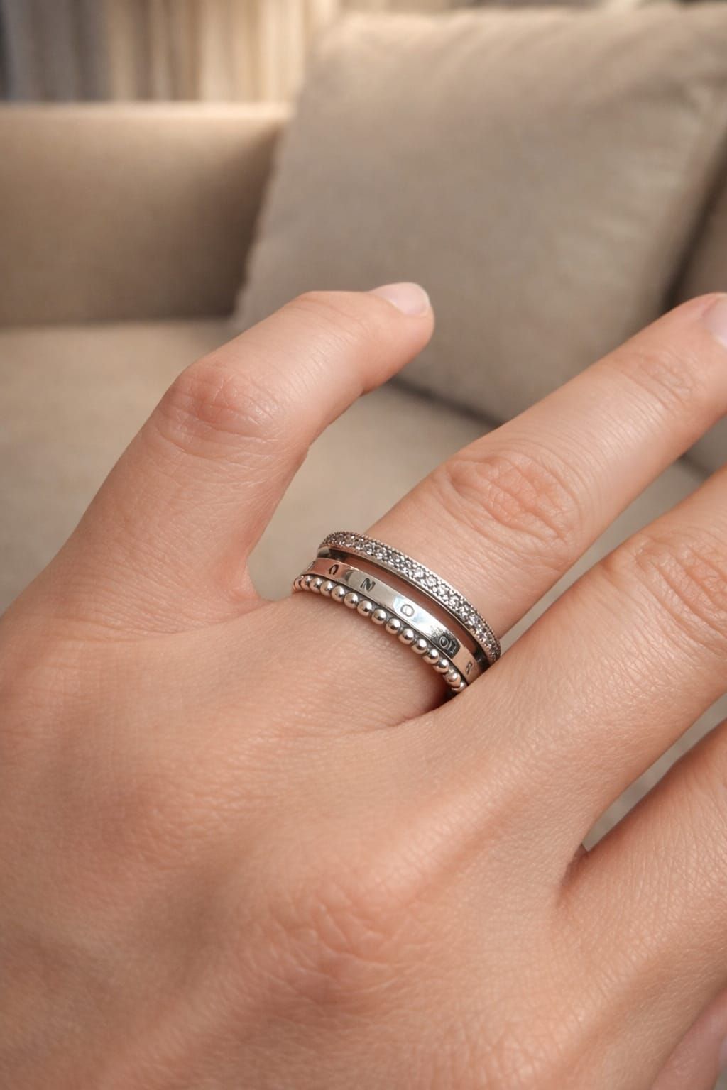 Una mano con anillos apilados de plata con pequeñas piedras descansa sobre un sofá beige.