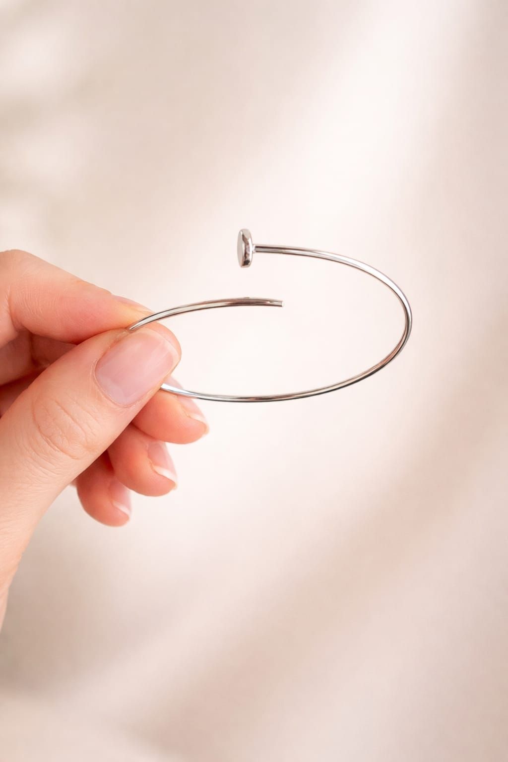 Mano sosteniendo una pulsera de plata abierta sobre un fondo beige suave.