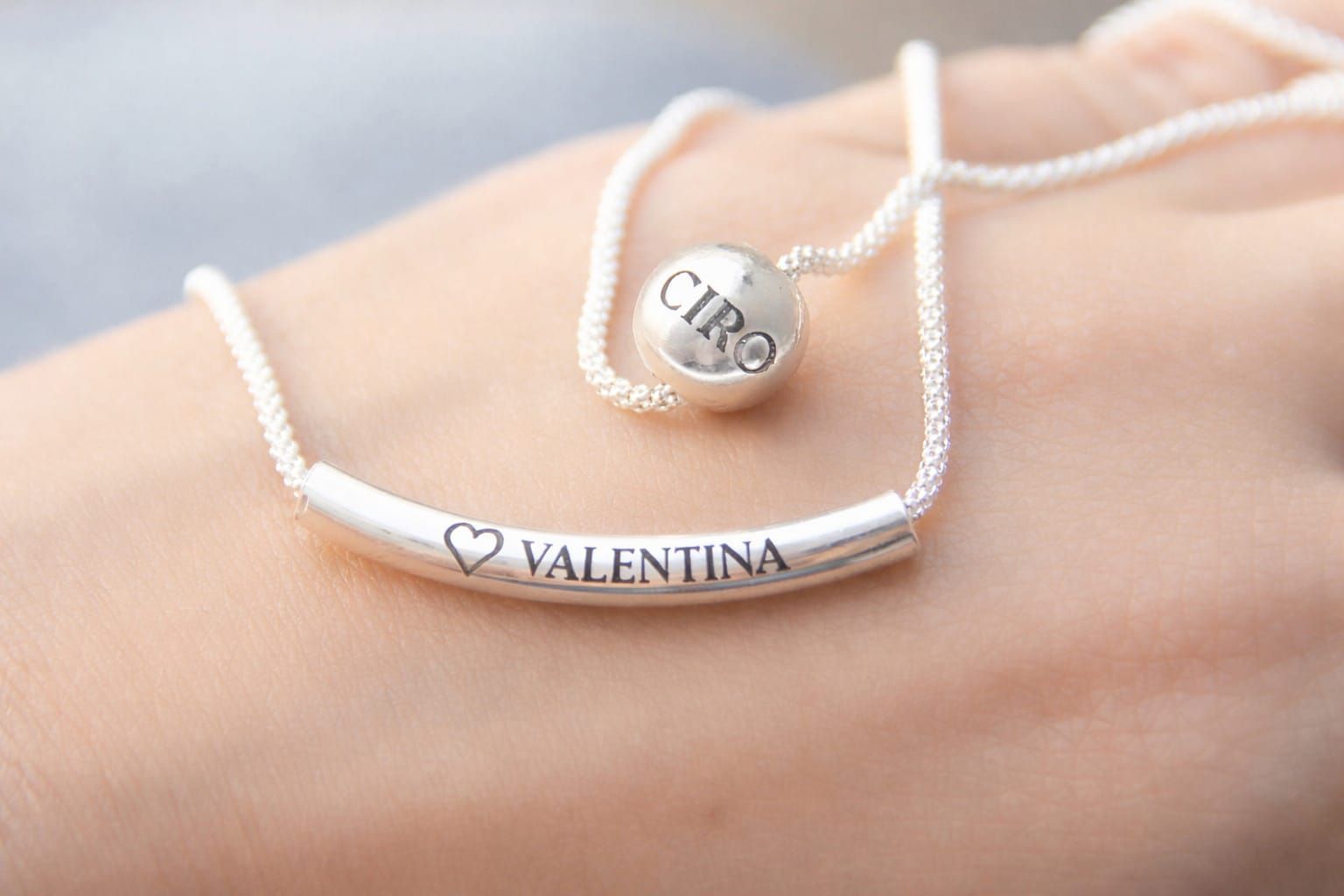 Collares de plata personalizados en una mano, con colgantes en forma de corazón y los nombres “Ciro” y “Valentina”.