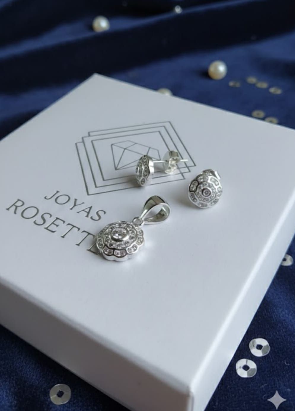Pendientes de plata en una caja blanca de JOYAS ROSETTE con un logotipo geométrico sobre fondo azul marino.