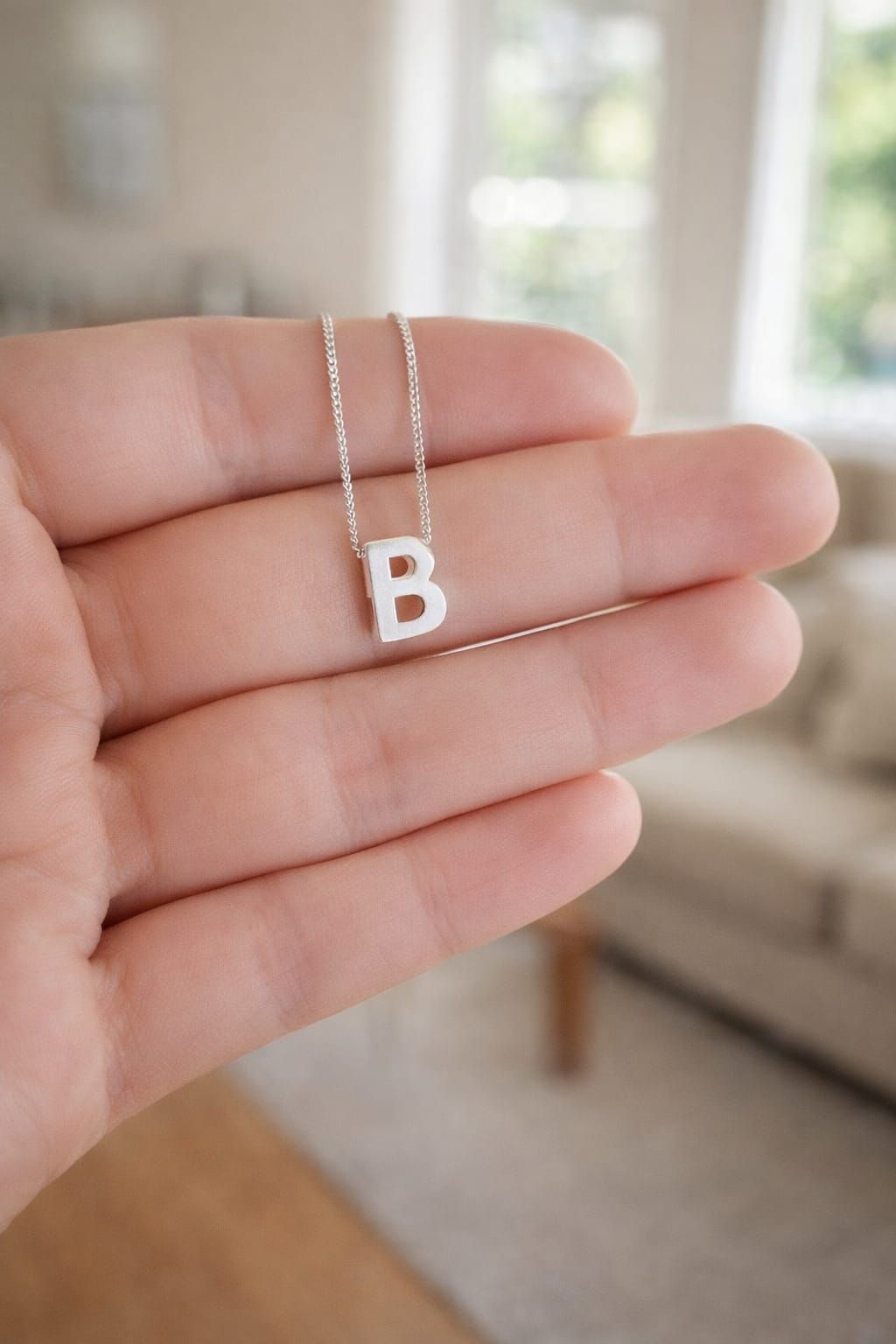 Mano sosteniendo un collar de plata con un colgante con la letra B sobre un fondo borroso de una sala de estar.
