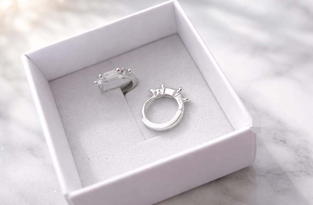 Una mano sostiene dos monturas de anillos de plata con pequeñas piedras preciosas sobre un fondo beige difuminado.