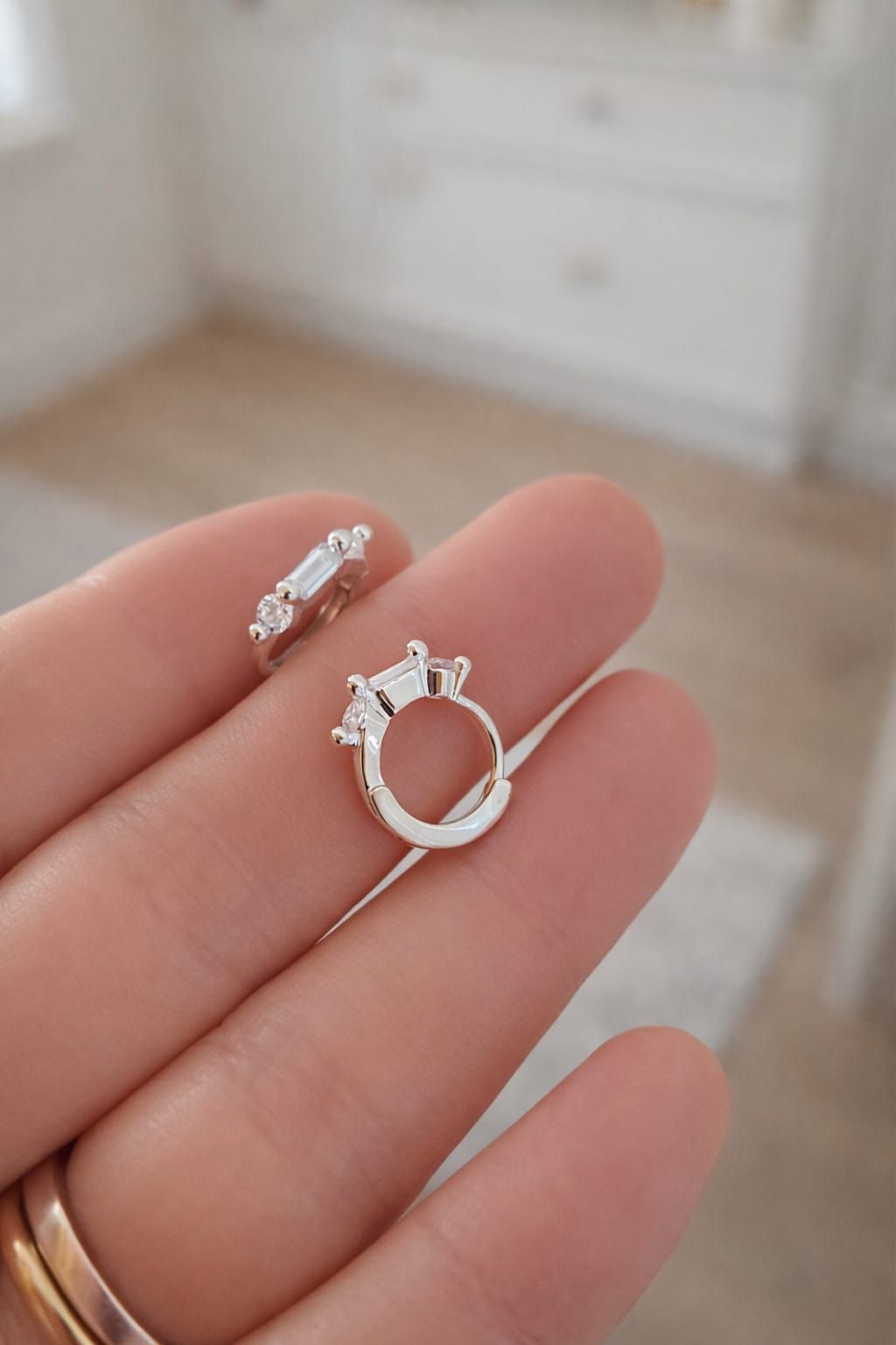 Una mano sostiene dos monturas de anillos de plata con pequeñas piedras preciosas sobre un fondo beige difuminado.