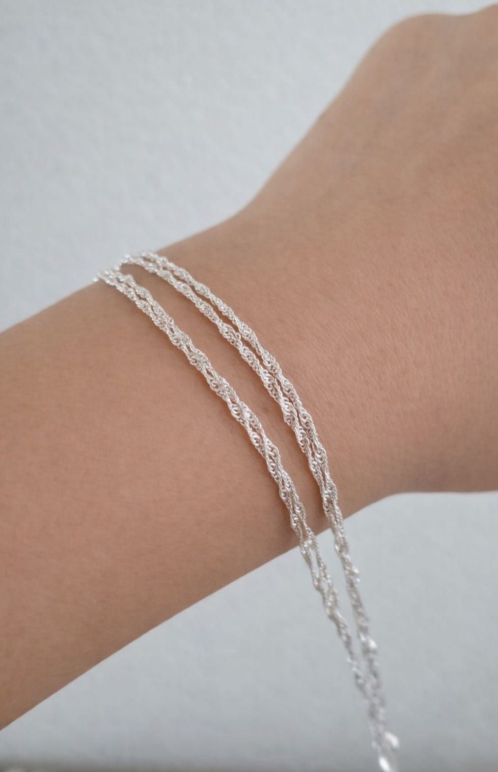 Una mano con tres delicadas pulseras de cadena de plata sobre un fondo claro.