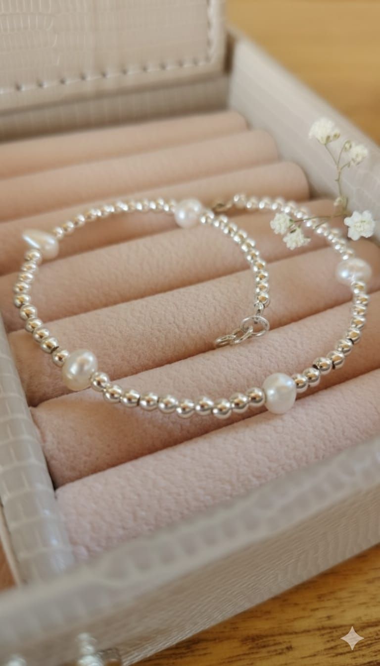 Una mano que luce una delicada pulsera de plata con un detalle de perla en una sala de estar suavemente desenfocada.