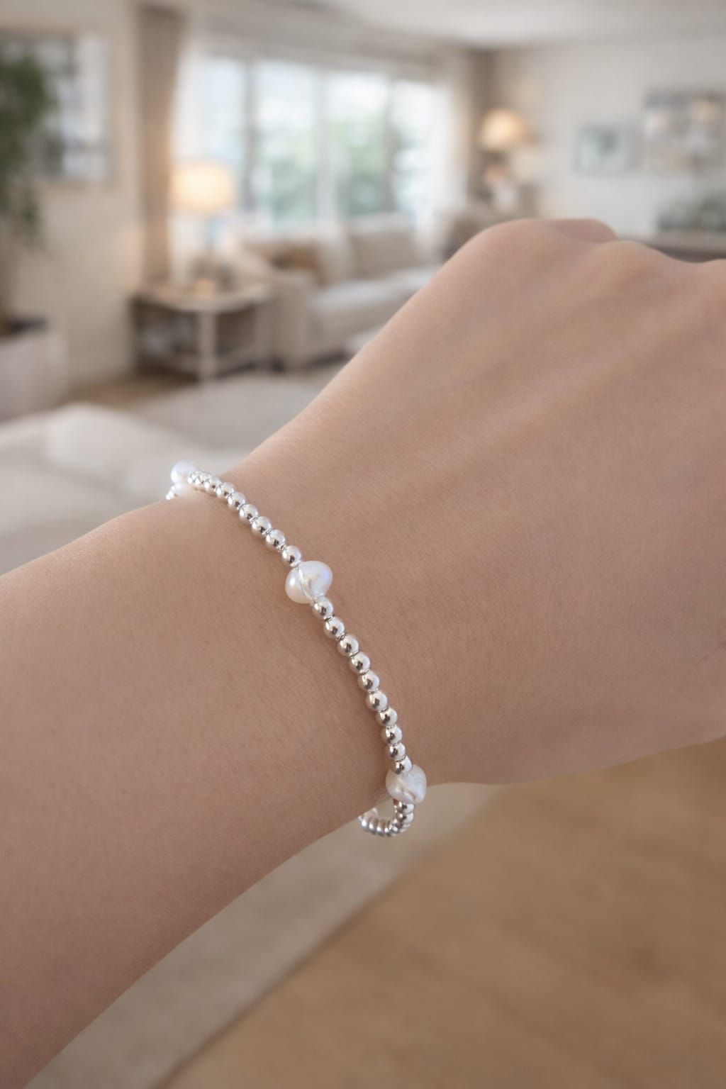 Una mano que luce una delicada pulsera de plata con un detalle de perla en una sala de estar suavemente desenfocada.