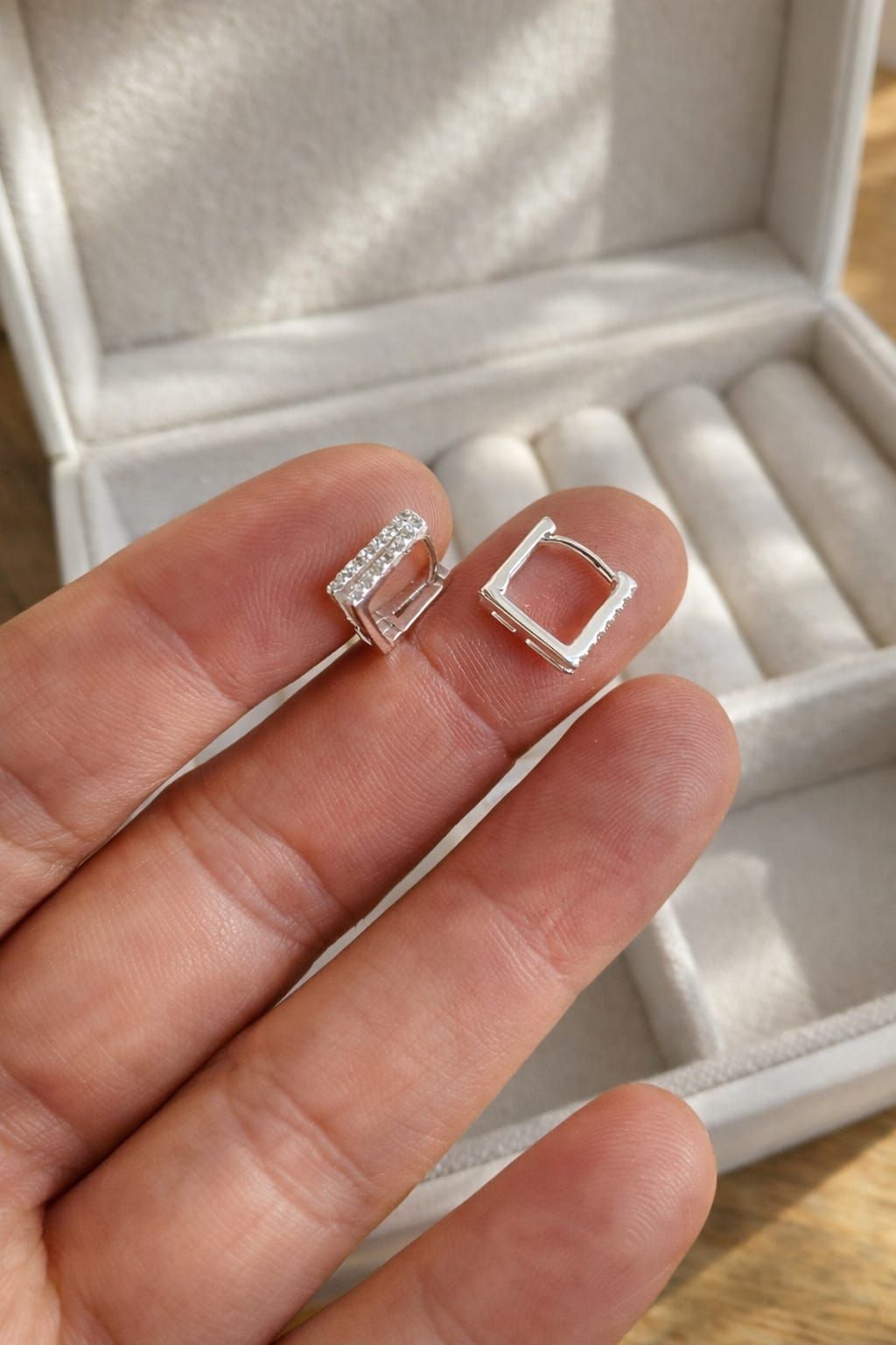 Dos pequeños pendientes cuadrados de plata sujetos entre los dedos, uno de ellos adornado con piedras transparentes.