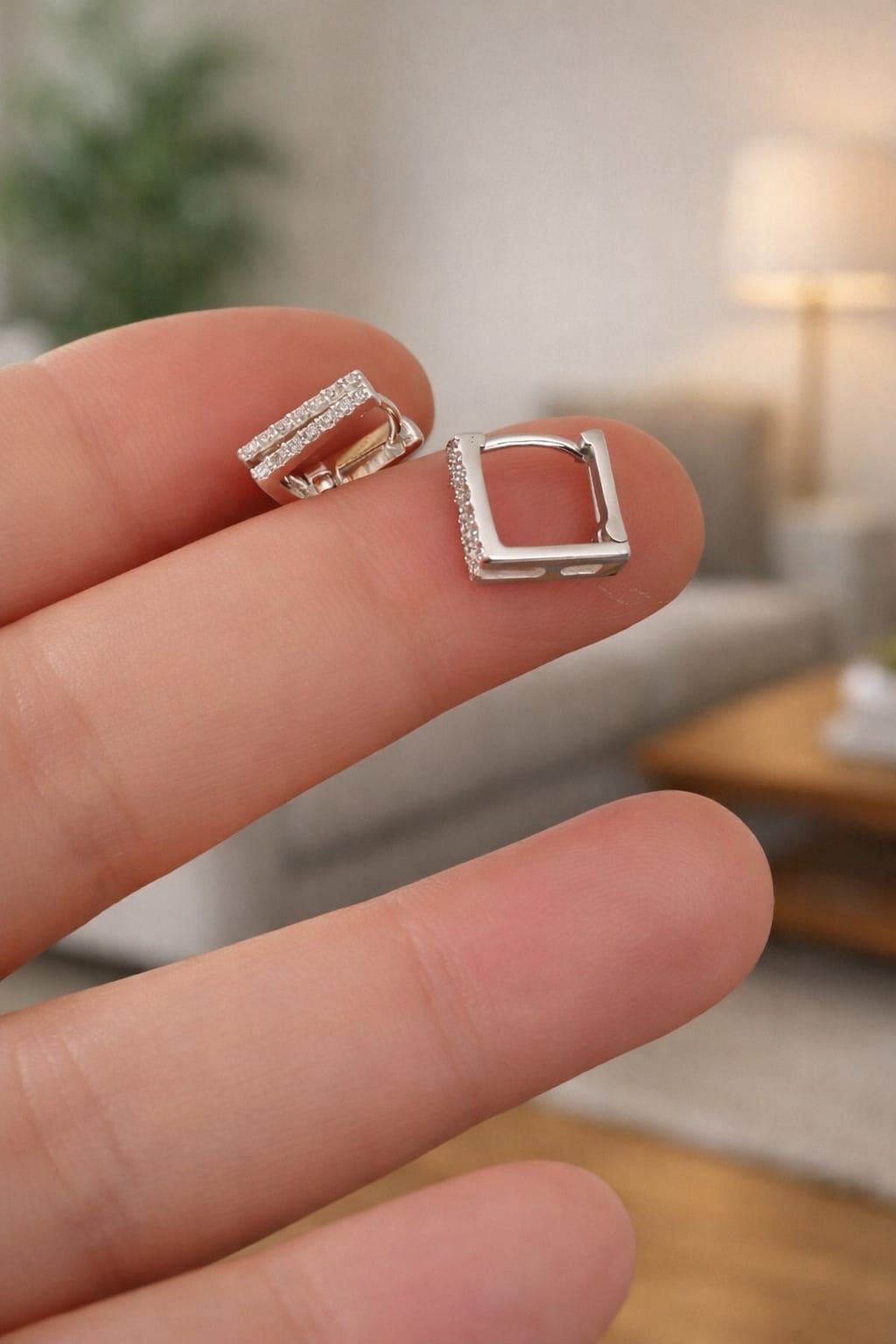Dos pequeños pendientes cuadrados de plata sujetos entre los dedos, uno de ellos adornado con piedras transparentes.