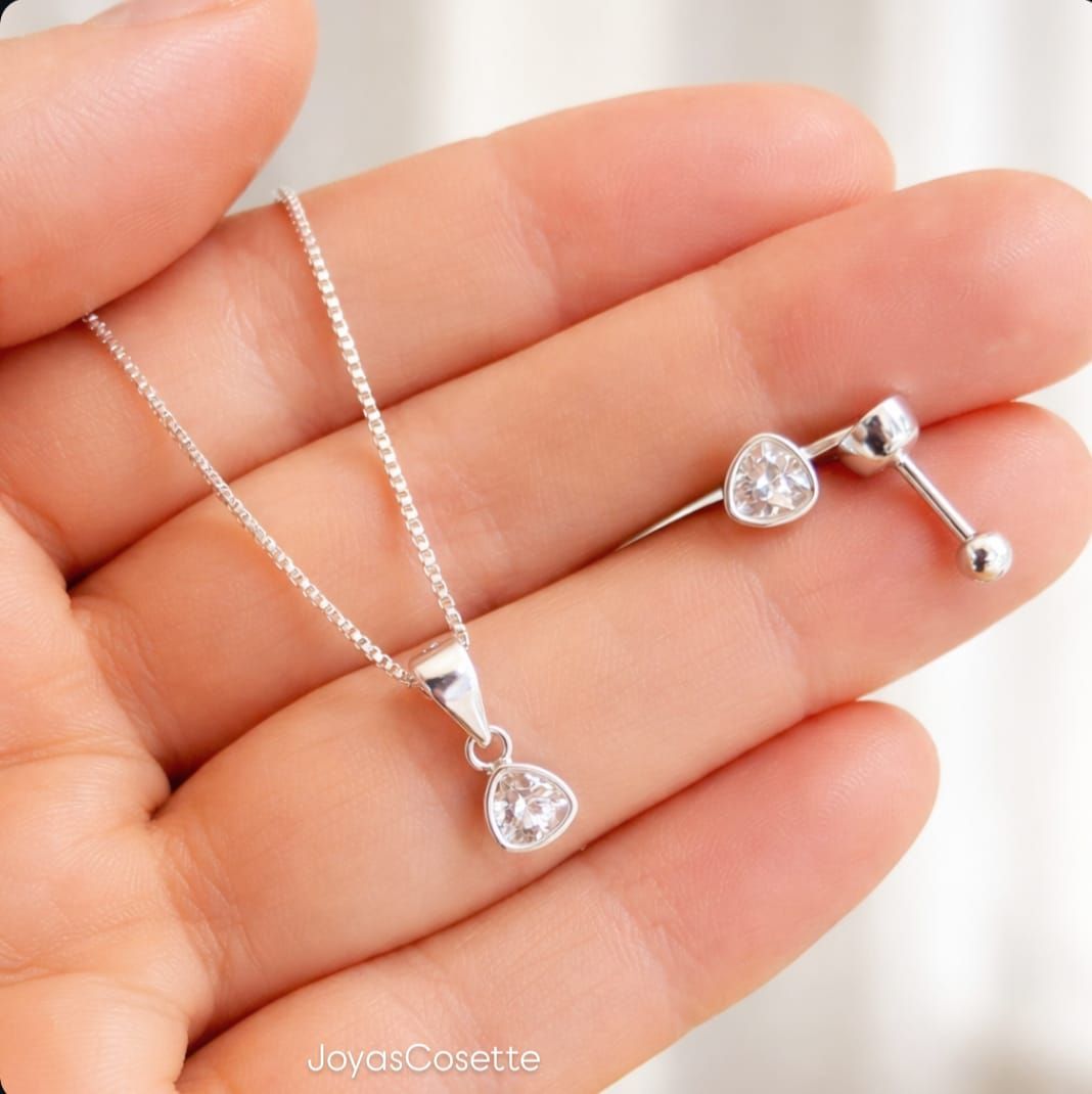 Collar con colgante de corazón de plata y pendientes a juego sostenidos en una mano sobre un fondo blanco.