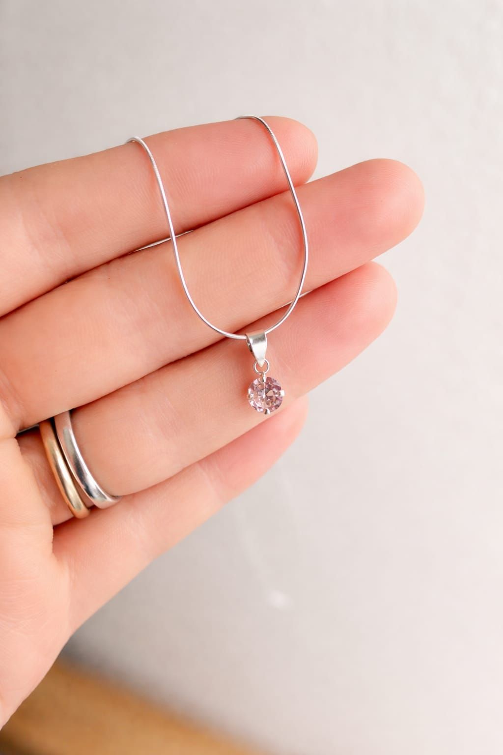 Mano sosteniendo un collar de plata con un pequeño colgante transparente sobre un fondo claro.