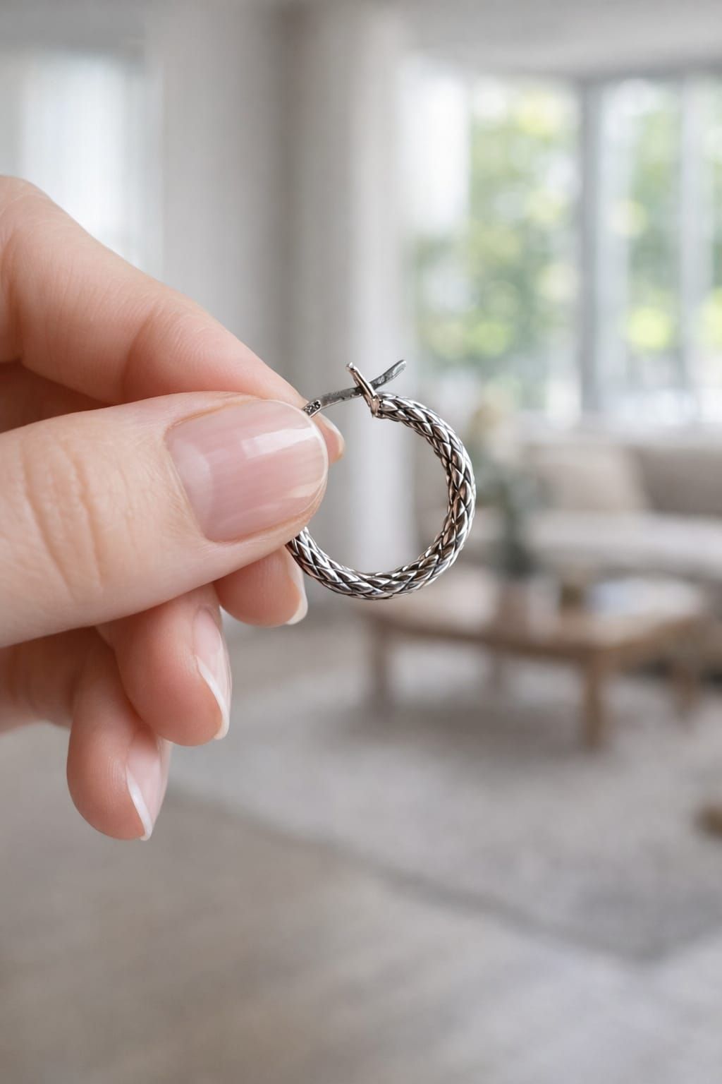 Una mano sostiene un pequeño pendiente de aro de plata con una piedra transparente en una luminosa sala de estar.