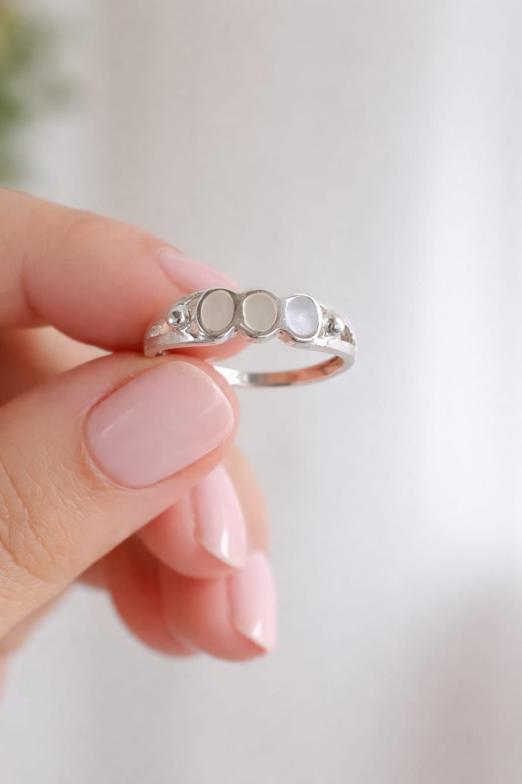 Mano sosteniendo un anillo de plata con pequeñas piedras transparentes sobre un fondo blanco suave.