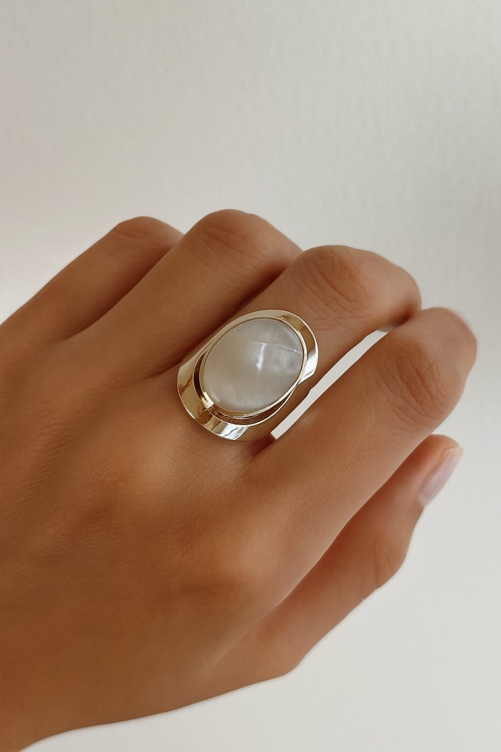 Mano con un gran anillo ovalado de piedra blanca sobre un fondo claro liso.