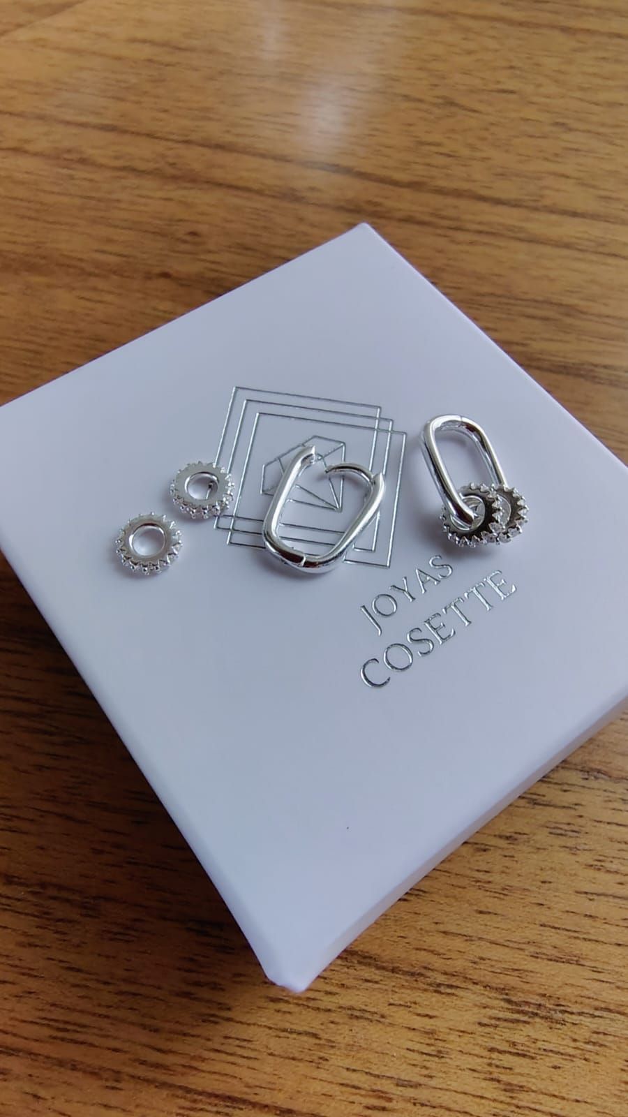 Dos pendientes colgantes de plata sostenidos entre los dedos sobre un fondo de tela rosa suave.