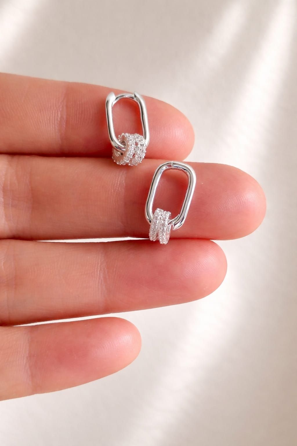 Dos pendientes colgantes de plata sostenidos entre los dedos sobre un fondo de tela rosa suave.