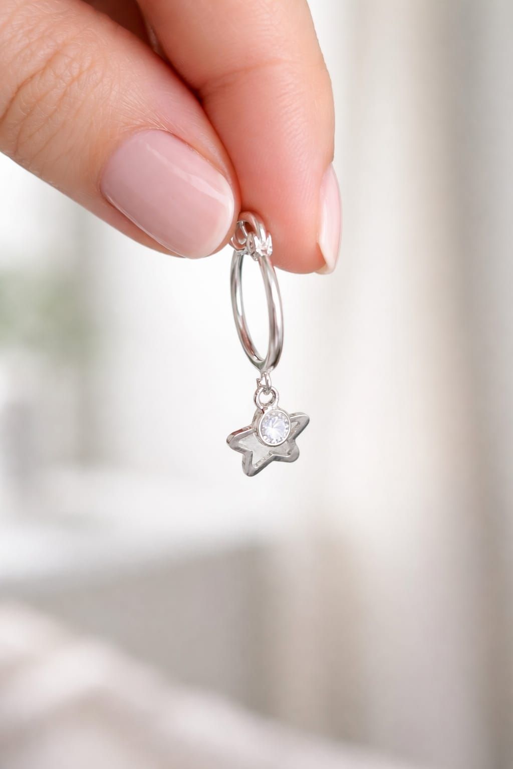 Mano sosteniendo un pendiente de aro plateado con un pequeño dije de estrella sobre un fondo suave.
