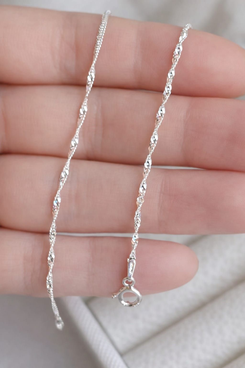 Un delicado collar de cadena de plata cuelga de una mano, con un pequeño broche en el extremo.