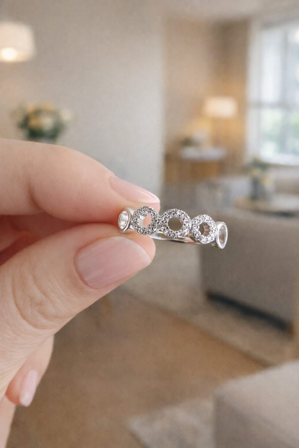 Mano sosteniendo un anillo de diamantes plateado con diseño de infinito sobre un fondo borroso de una sala de estar.