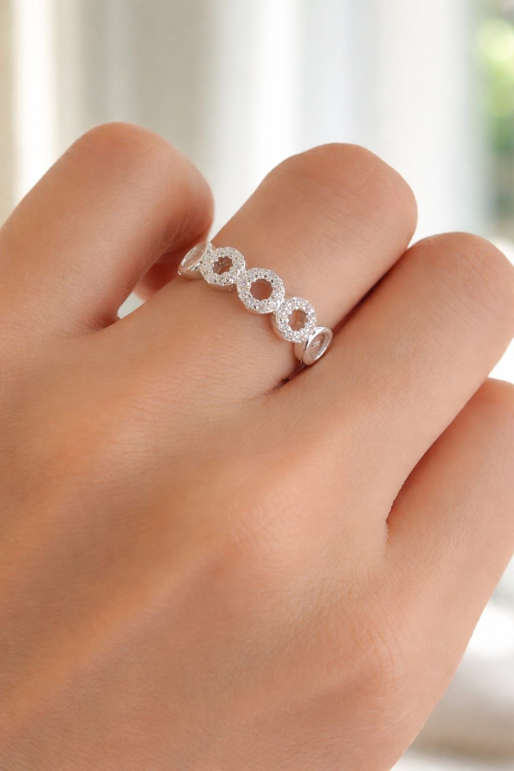 Mano sosteniendo un anillo de diamantes plateado con diseño de infinito sobre un fondo borroso de una sala de estar.