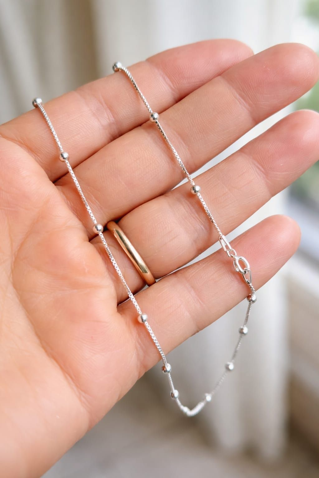 Mano sosteniendo un delicado collar de cadena de cuentas de plata sobre un fondo suave y desenfocado.