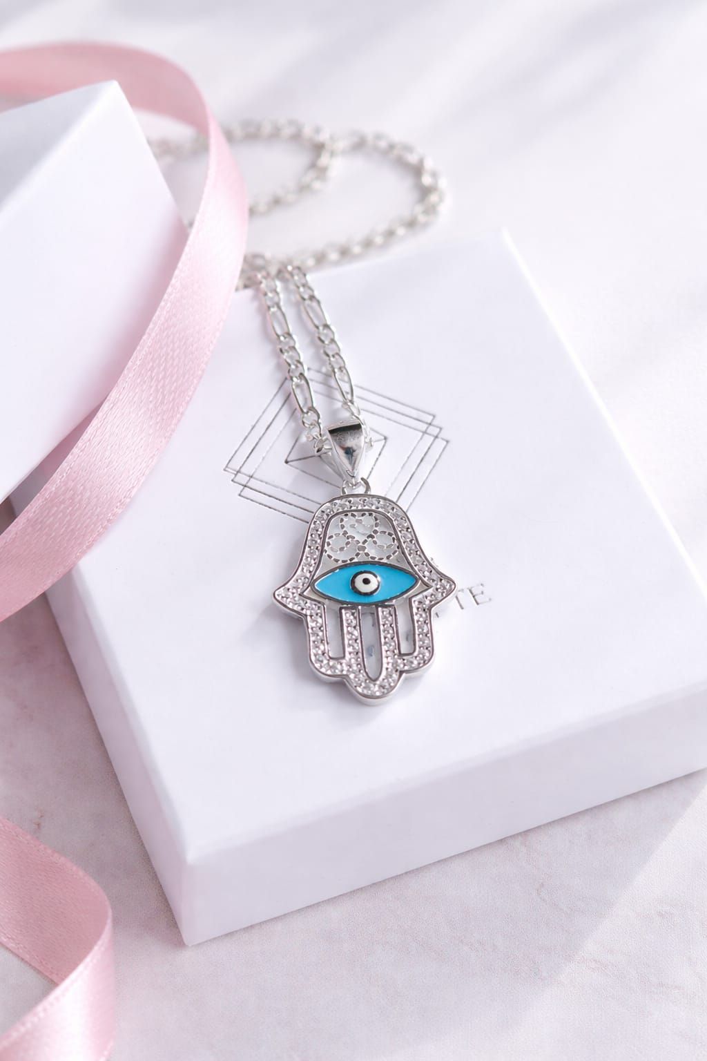 Collar con colgante Hamsa de plata con ojo azul en una caja de regalo rosa pálido y blanca.