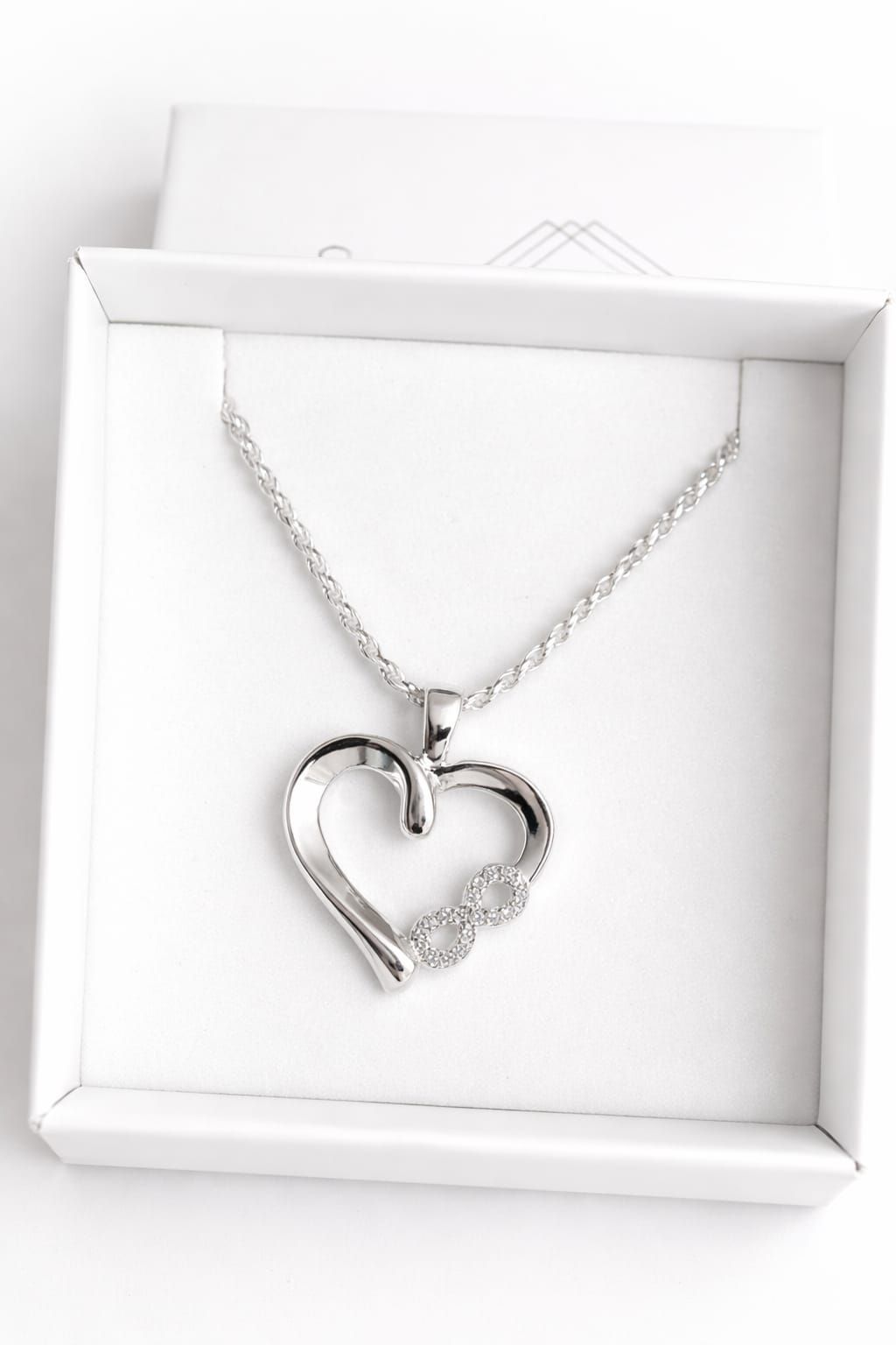Colgante de plata en forma de corazón con el símbolo del infinito, sostenido entre los dedos.
