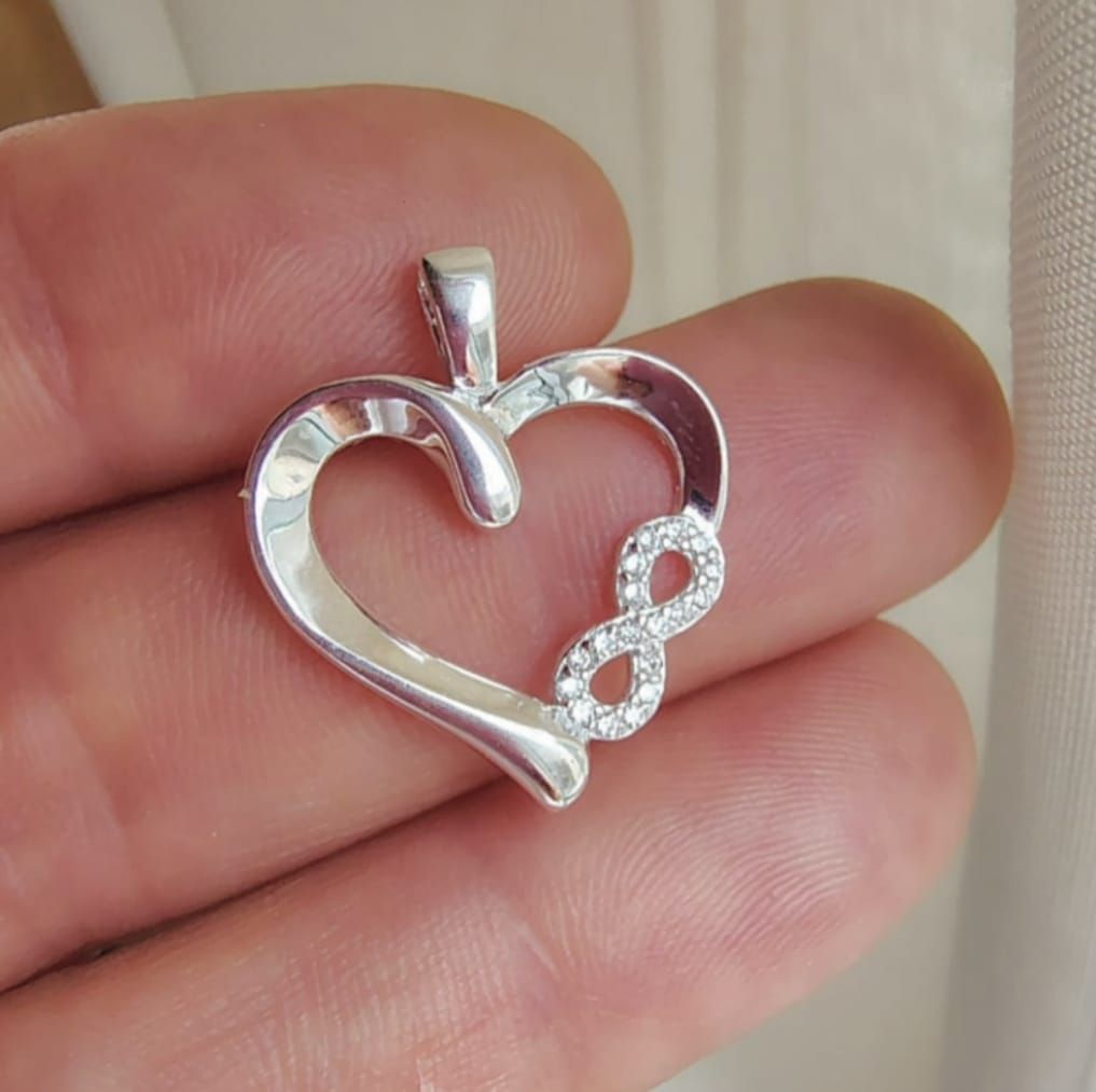 Colgante de plata en forma de corazón con el símbolo del infinito, sostenido entre los dedos.
