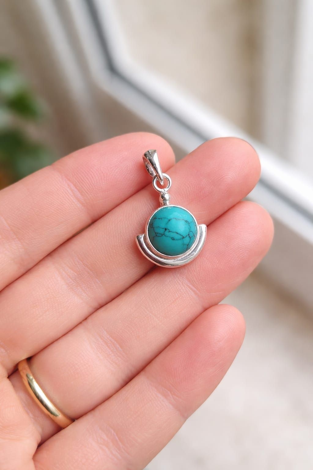 Mano sosteniendo un pequeño colgante de plata con una piedra azul turquesa, cerca de una ventana.