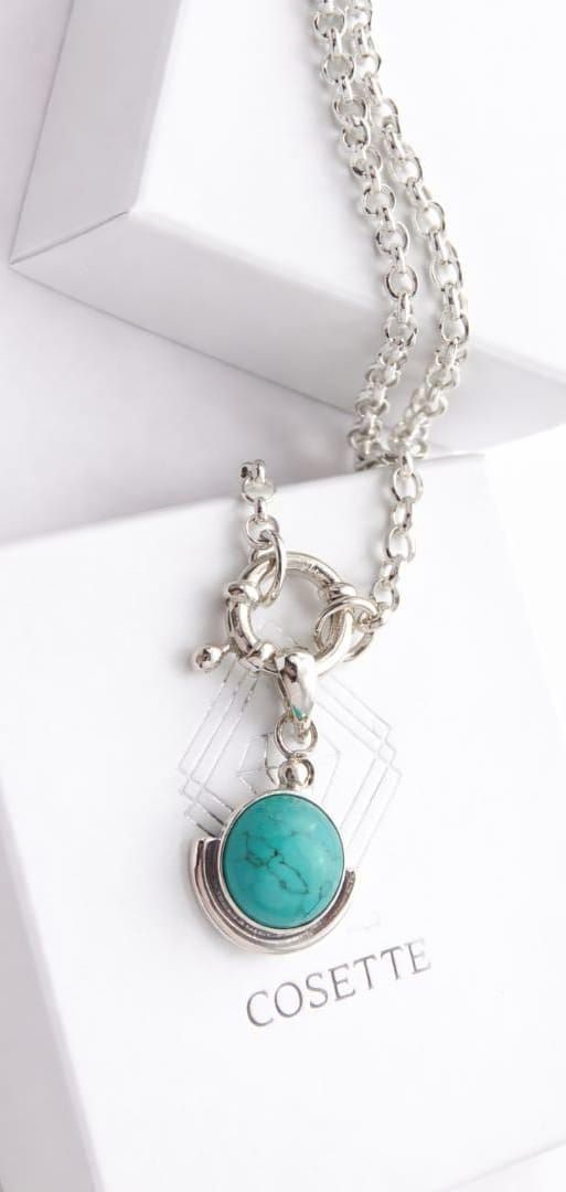 Mano sosteniendo un pequeño colgante de plata con una piedra azul turquesa, cerca de una ventana.