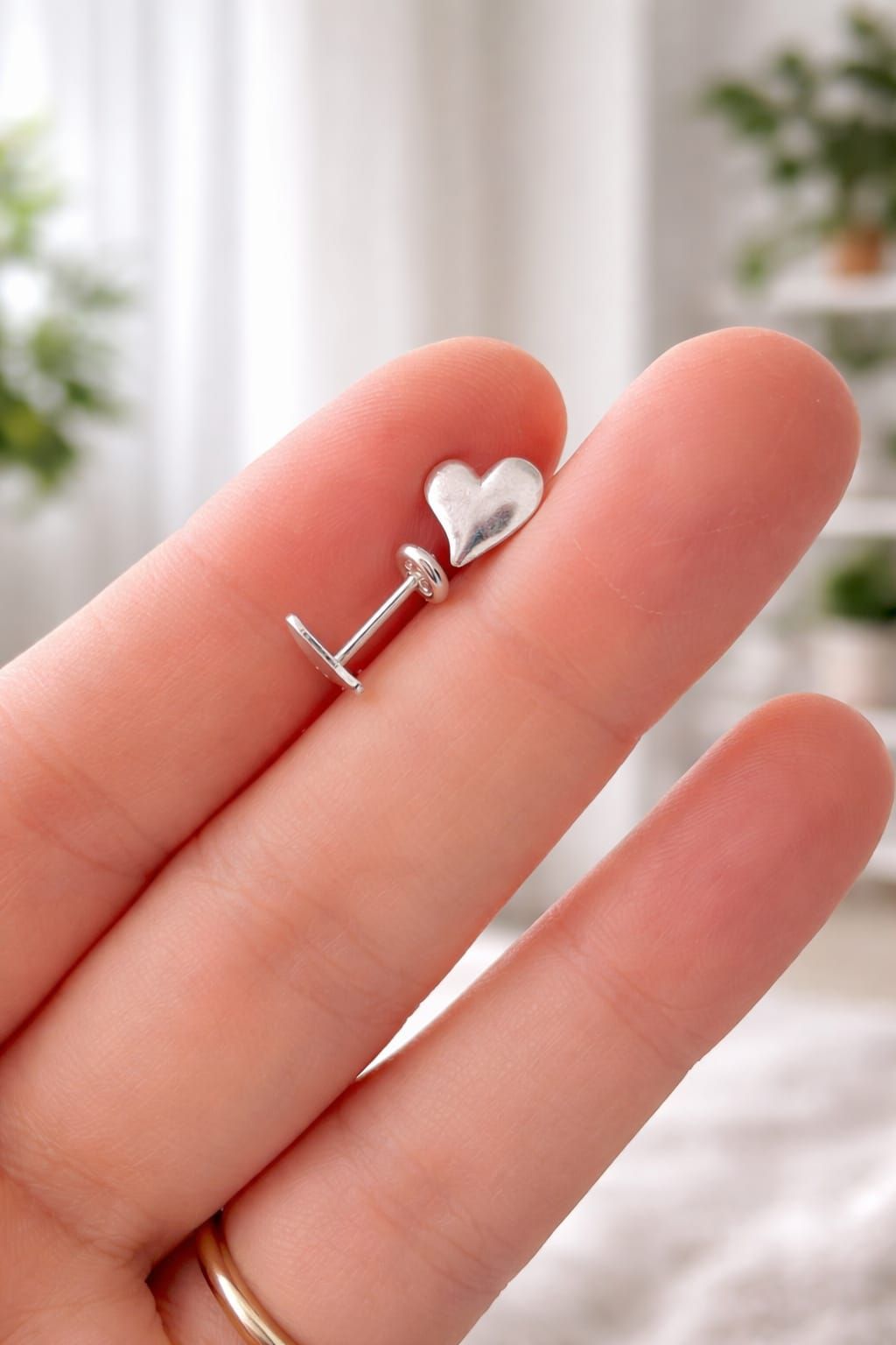 Pendiente pequeño de plata con forma de corazón, sostenido entre los dedos, con un fondo interior borroso.