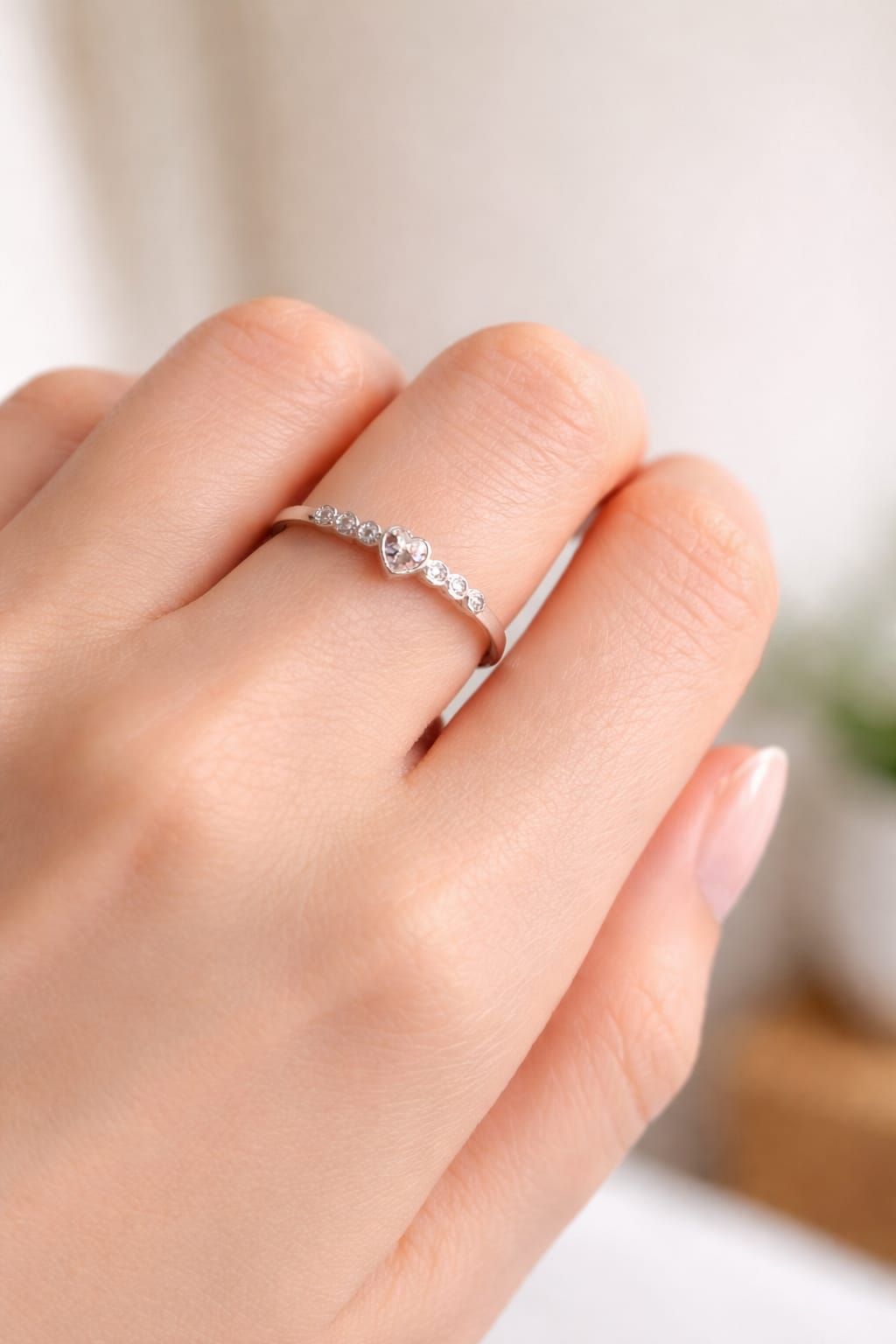 Primer plano de una mano que luce un delicado anillo de plata con pequeñas piedras sobre un fondo rosa pálido.