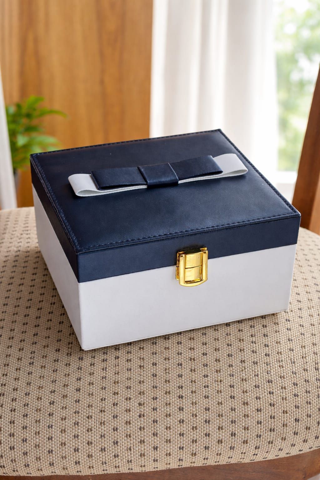 Pequeña caja decorativa en blanco y negro con cierre dorado sobre una mesa de mimbre cerca de una ventana.