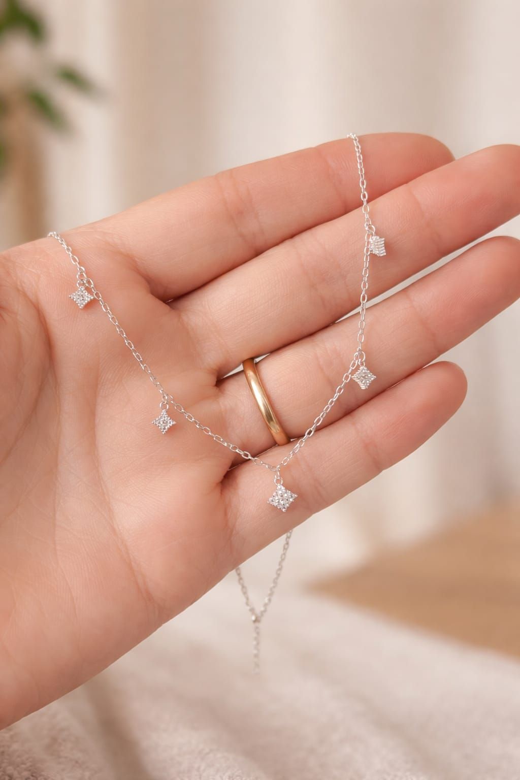 Mano sosteniendo un delicado collar de plata con pequeños detalles de cristal sobre un fondo beige suave.