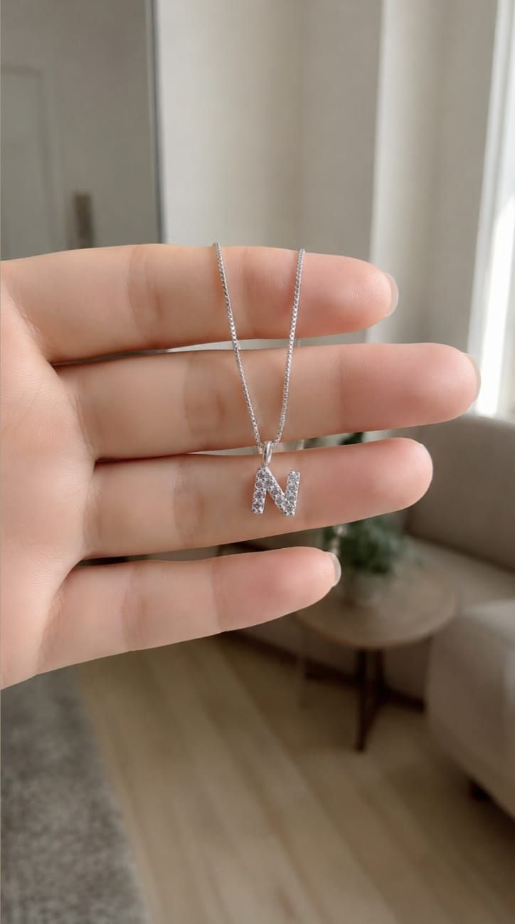 Una mano sostiene un collar de plata con un colgante brillante con la letra M en el interior de una vivienda.