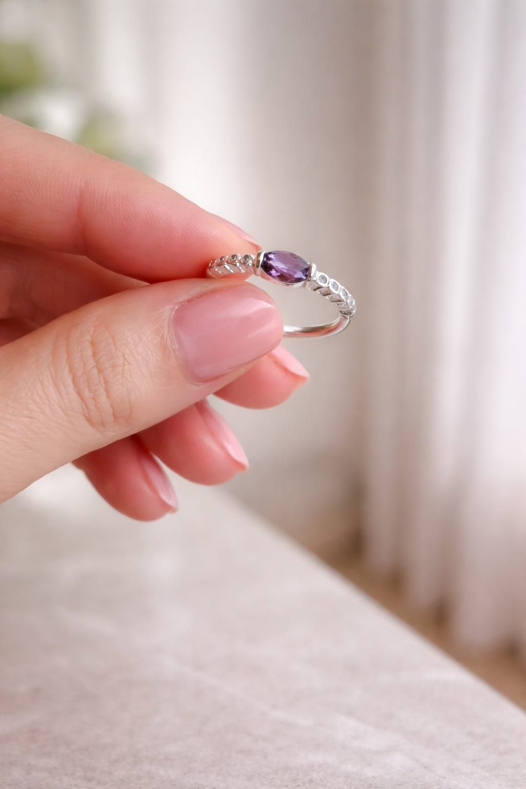 Mano con un anillo de plata con una gema púrpura sobre un fondo beige suave.
