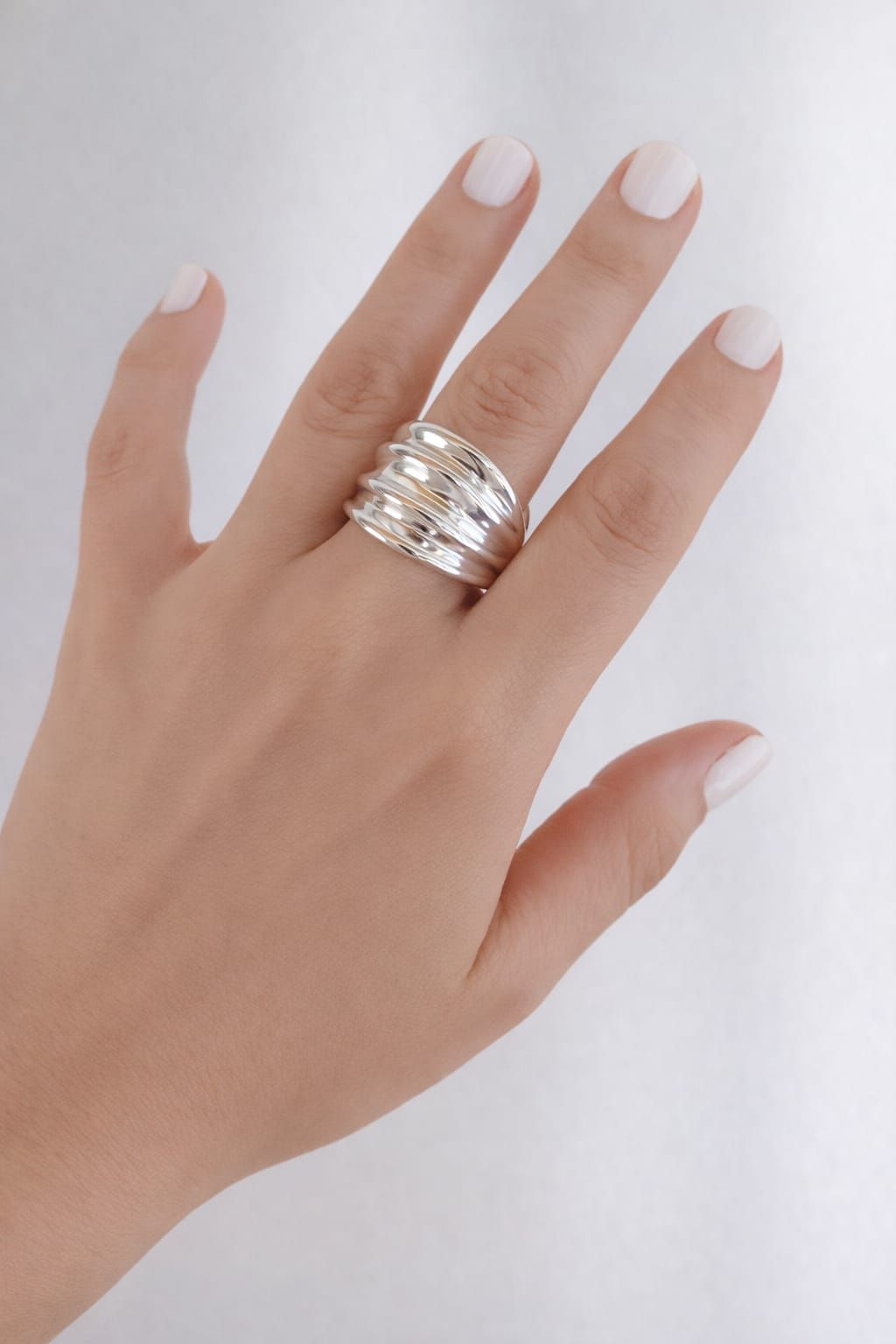 Mano con uñas blancas luciendo un anillo plateado apilado sobre un fondo blanco.