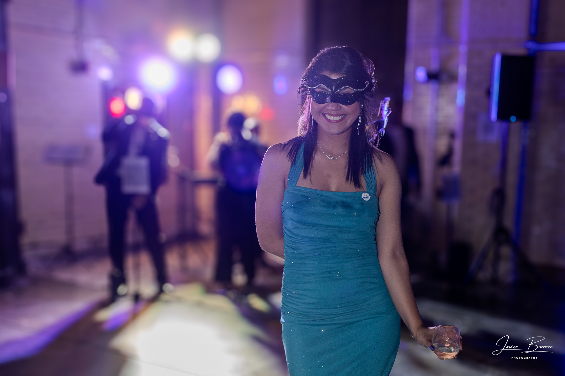 Woman in a teal dress and masquerade mask holding a drink at a party with blurred lights in the background