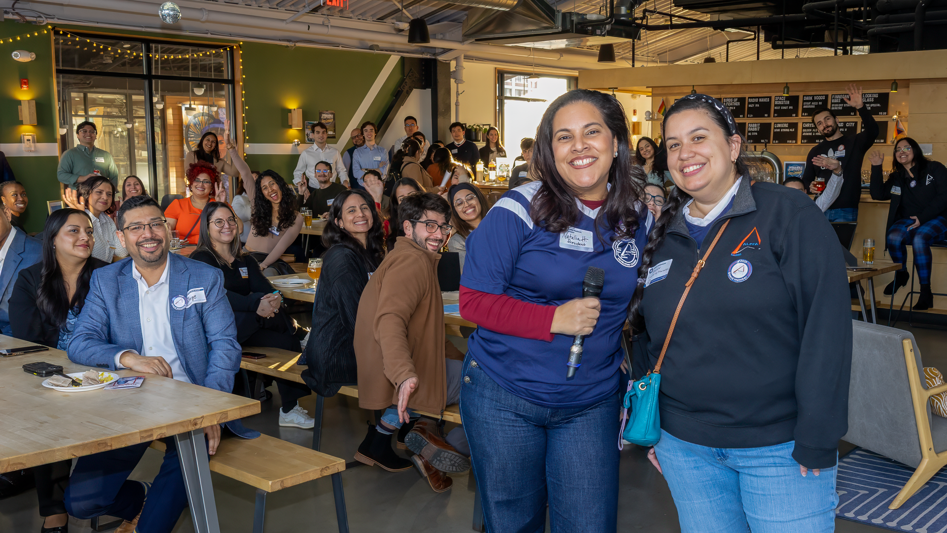 Two women smile while holding a microphone at an indoor social gathering with seated guests in a modern, casual venue.