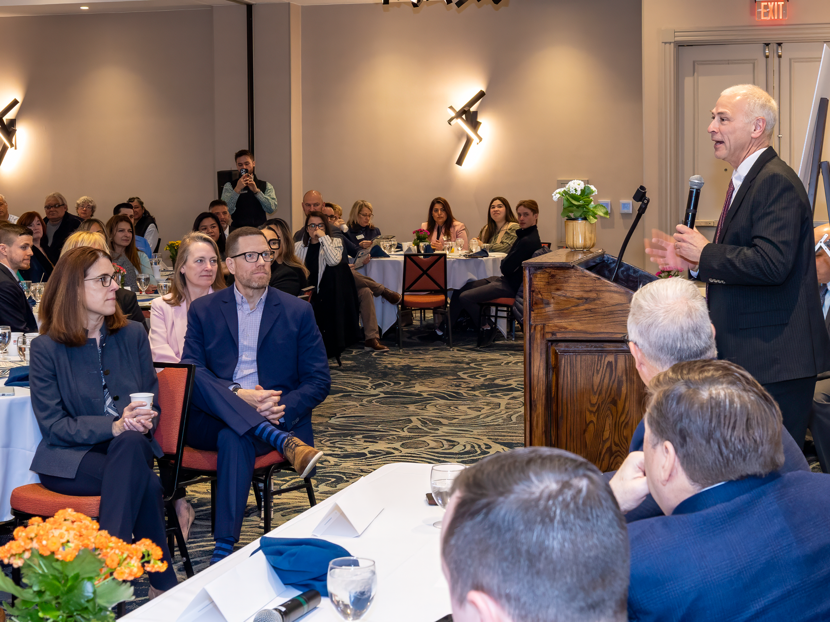 A speaker at a podium addresses a seated audience in a conference room with tables and floral centerpieces.