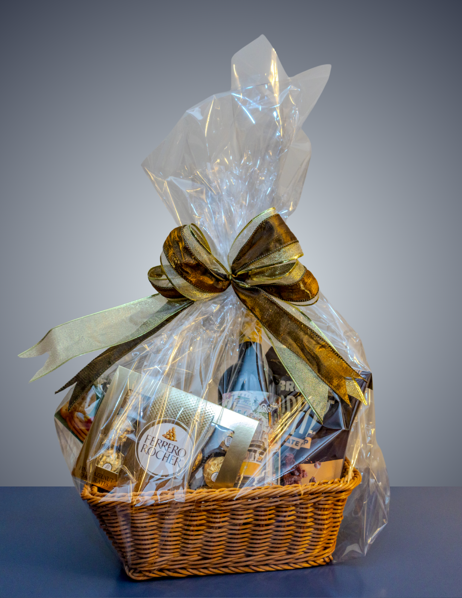 A wicker gift basket containing chocolates and a bottle, wrapped in clear cellophane with a large gold decorative bow.
