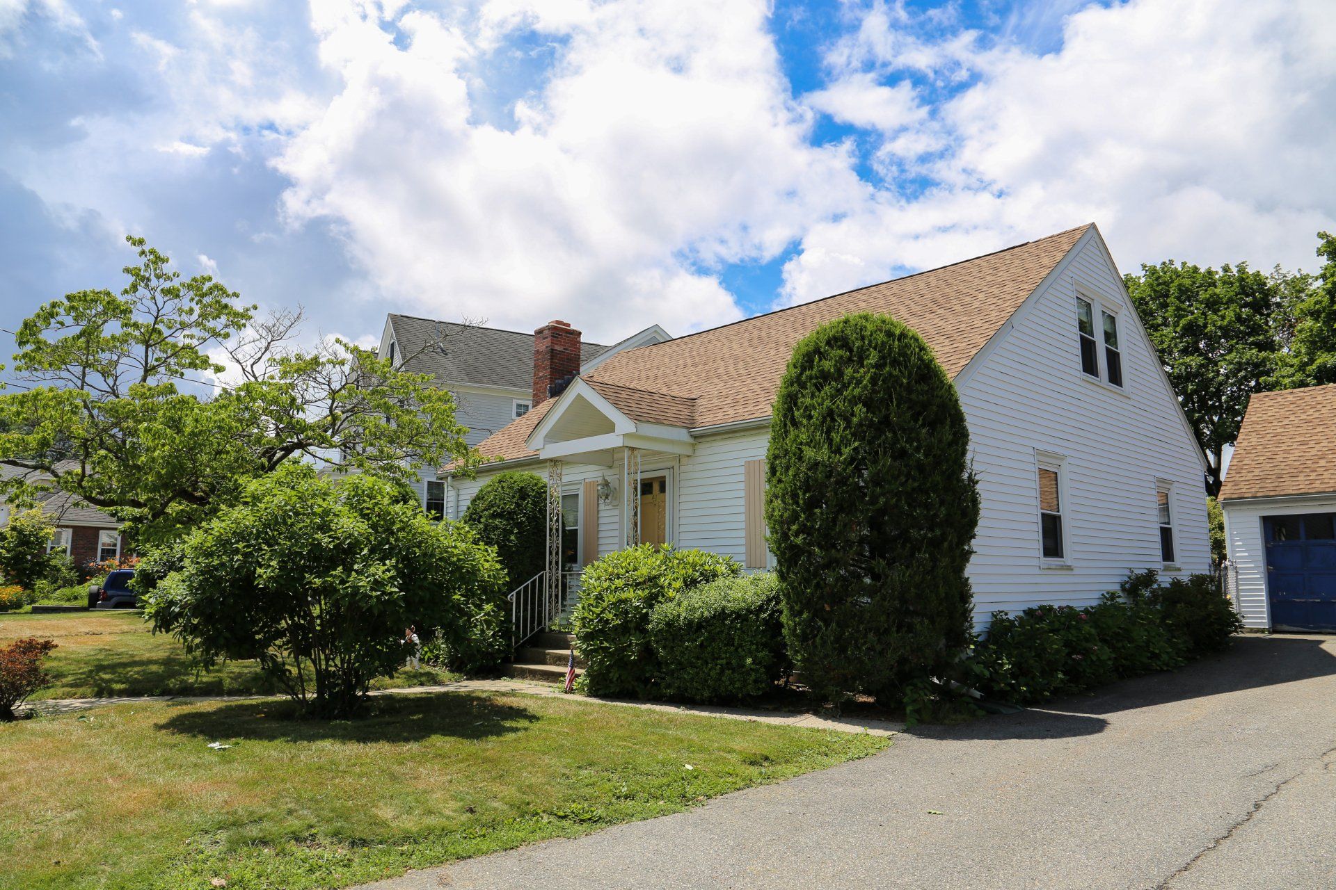 White house with brown roof, front gable entry, and manicured shrubs under a blue sky with clouds.