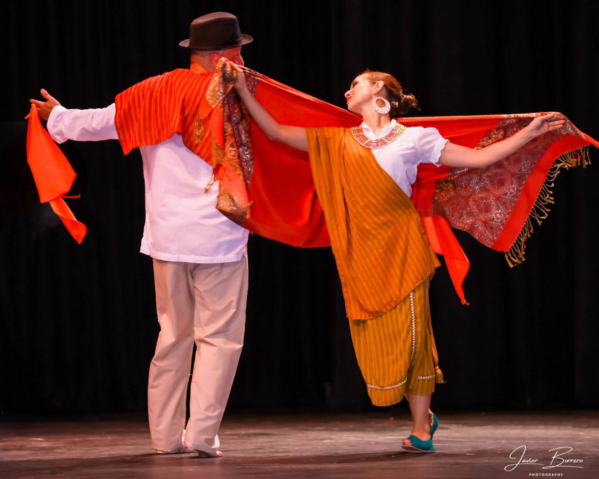 Two cultural dancers on stage connected by a red textile, wearing red and mustard traditional attire