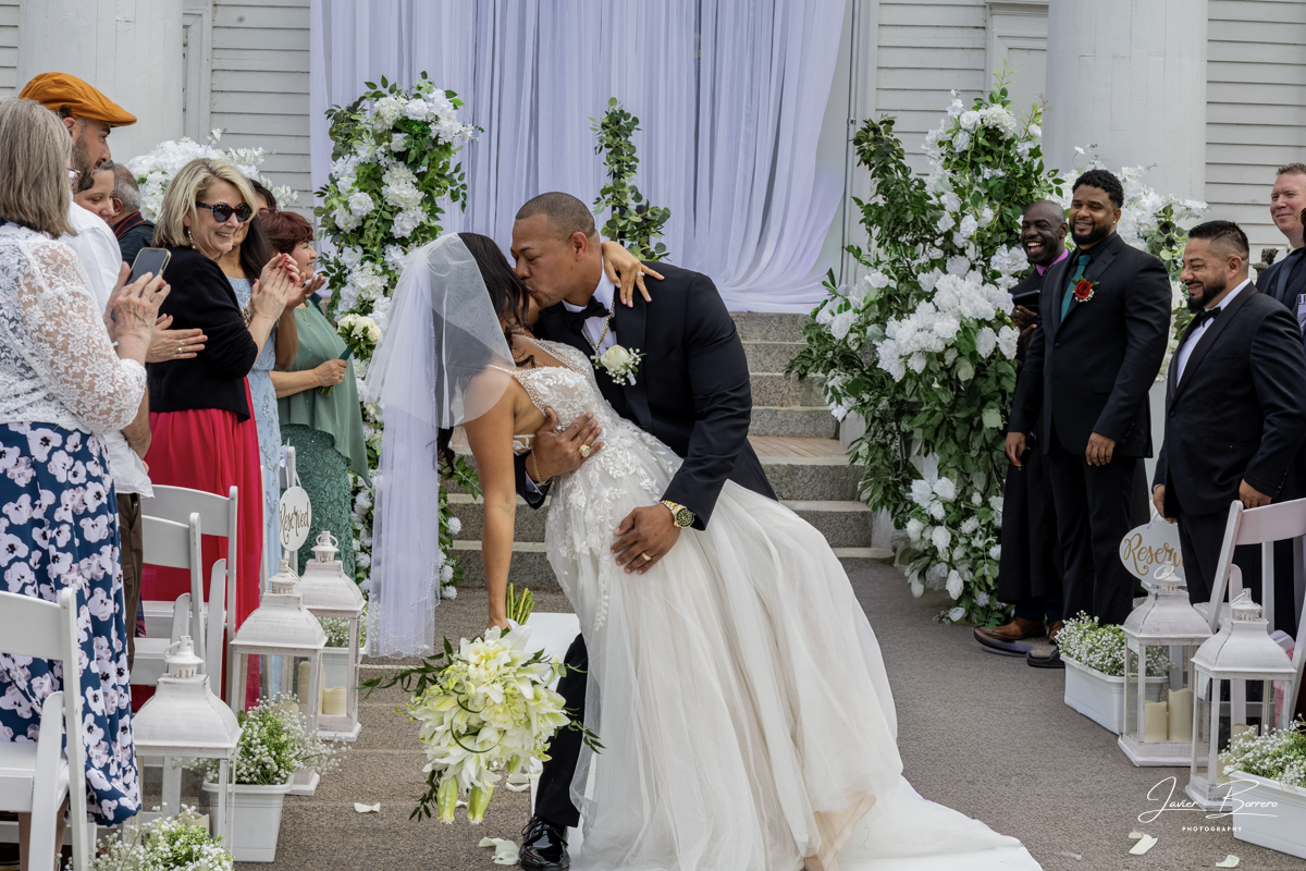 Wedding couple kissing at the altar in an outdoor ceremony, surrounded by white flowers and seated guests