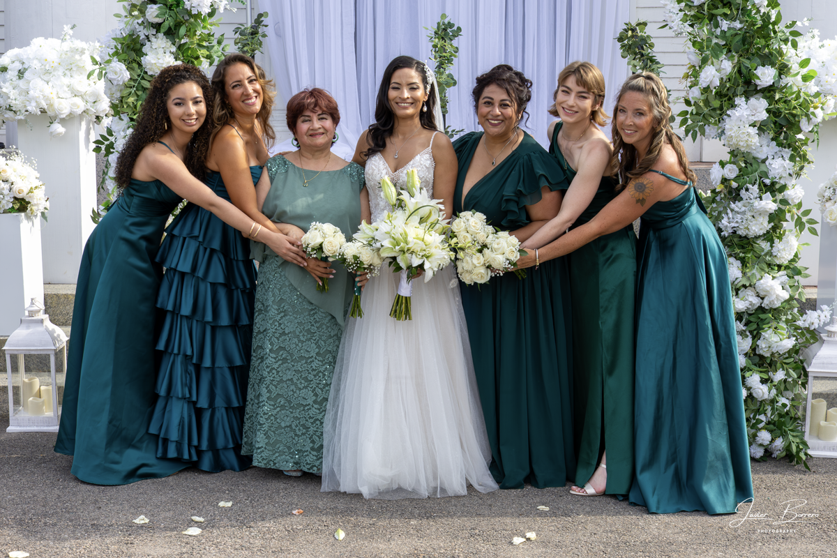 Wedding party in teal dresses posing with bouquets under a white floral arch outdoors