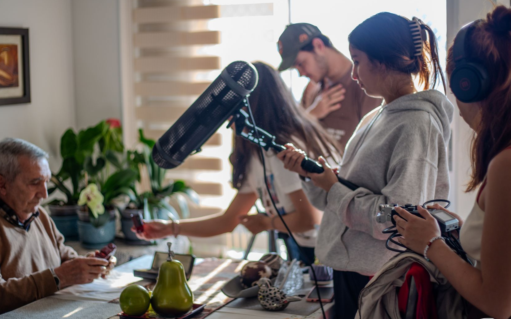 People recording a podcast at a kitchen table with a seated guest and microphones on stands